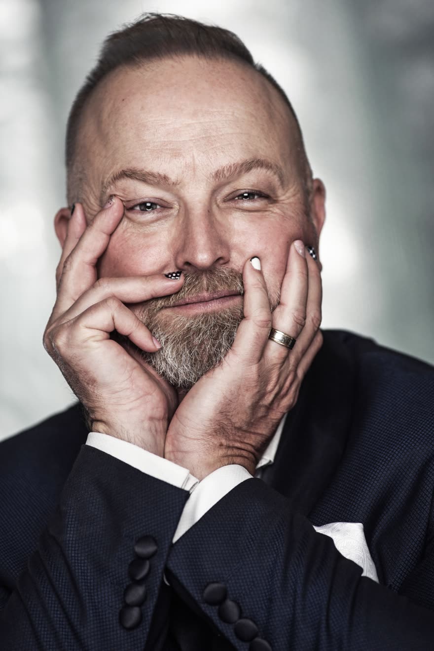 Portrait of Michael Bach: shaved head, salt-and-pepper beard, navy suit, hands framing the face, half-smiling at camera.